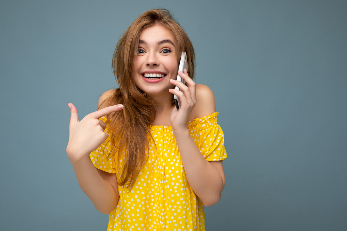 Photo of Beautiful Positive Surprised Young Blonde Woman Wearing Stylish Yellow Summer Dress Standing Isolated over Blue Background Holding and Talking on Mobile Phone with Sincere Emotions Looking at Camera Pointing Finger at Yourself